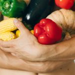 A man's torso holding a variety of fresh vegetables including peppers and corn.