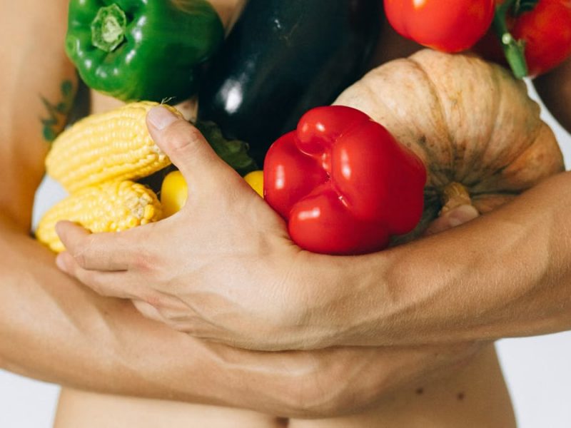 A man's torso holding a variety of fresh vegetables including peppers and corn.