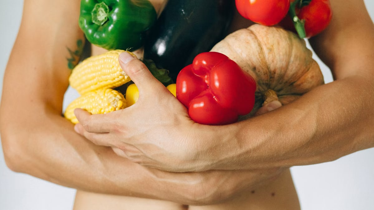 A man's torso holding a variety of fresh vegetables including peppers and corn.
