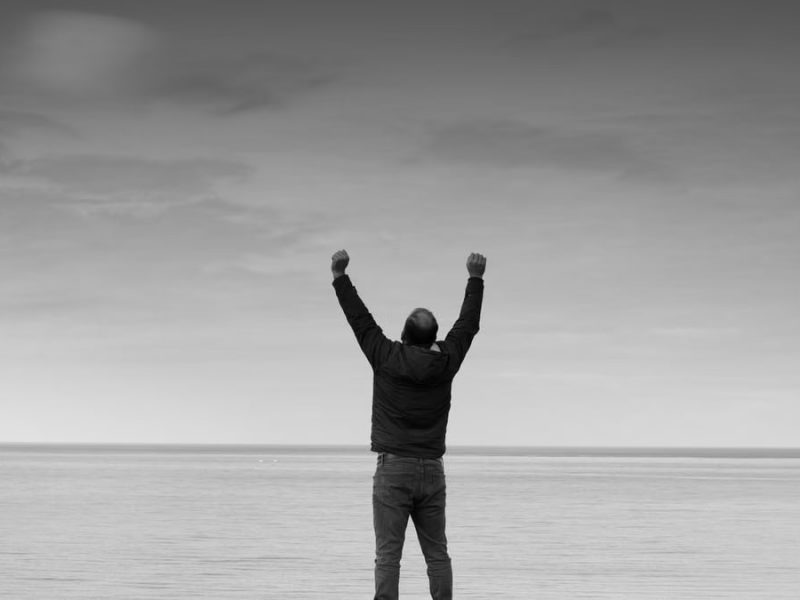 Black and white image of a person raising arms in celebration by the sea.